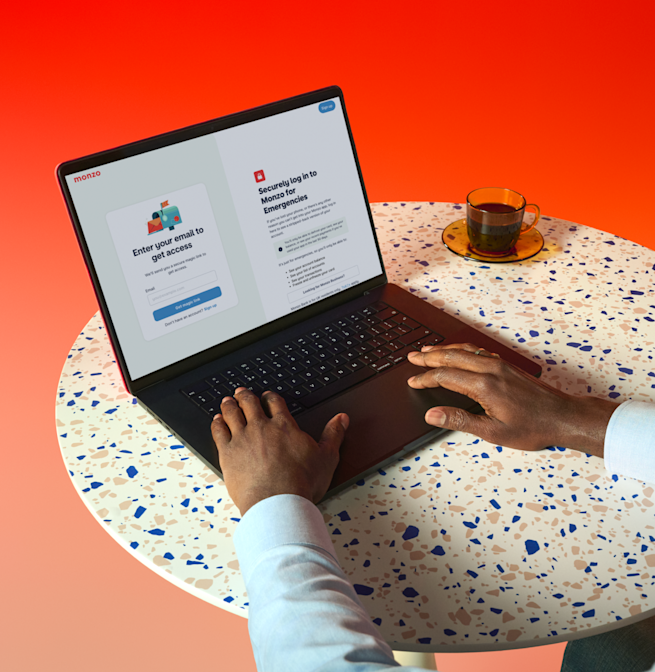 A high-angle shot shows a person's hands typing on a laptop displaying the Monzo banking app login page. The laptop is open on a round, light-colored table with multicolored specks. To the right of the laptop is a transparent teacup with a dark liquid, sitting on a matching saucer. The background is a vibrant gradient, transitioning from orange at the top to red at the bottom.