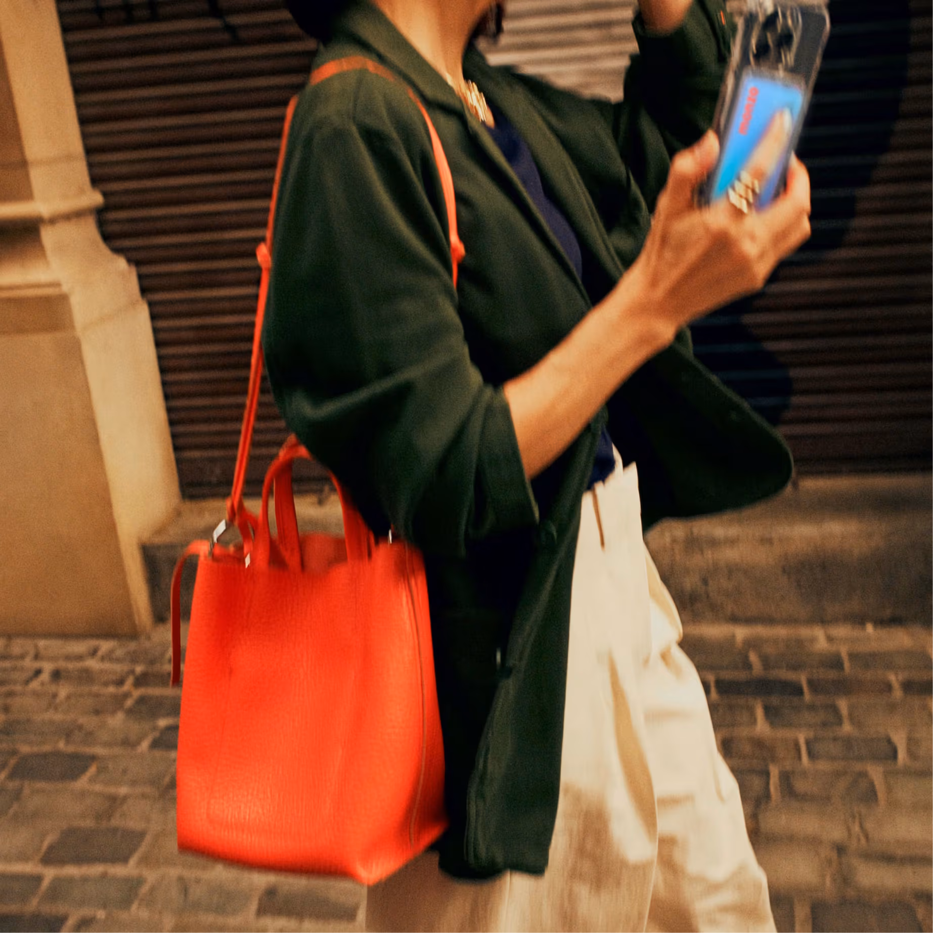 A person walks along a cobblestone street, wearing a dark green coat and cream trousers, carrying a hot coral coloured leather tote bag on their shoulder and holding a smartphone in their right hand. A Monzo debit card can be seen inside the phone case.