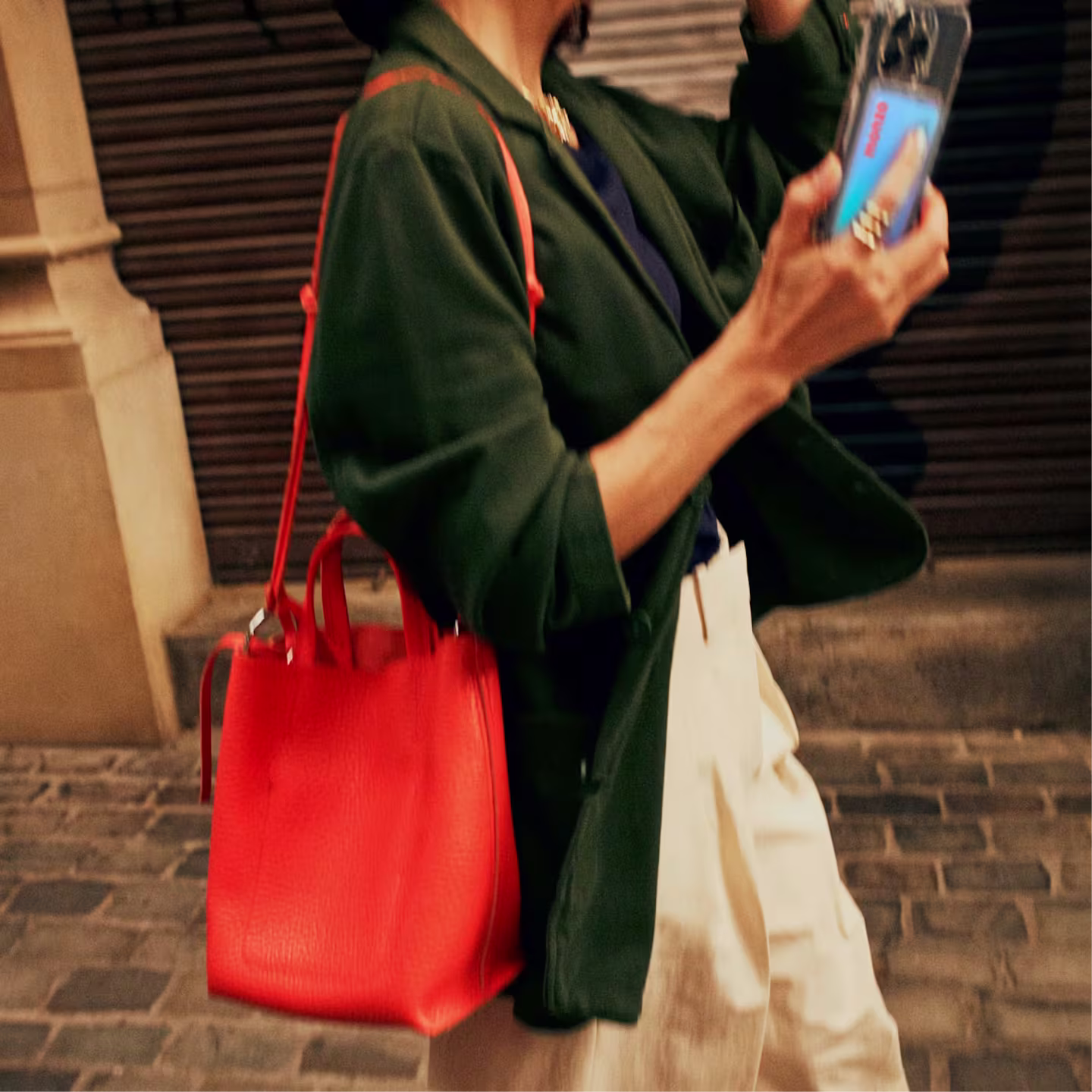 A person walks along a cobblestone street, wearing a dark green coat and cream trousers, carrying a hot coral coloured leather tote bag on their shoulder and holding a smartphone in their right hand. A Monzo debit card can be seen inside the phone case.