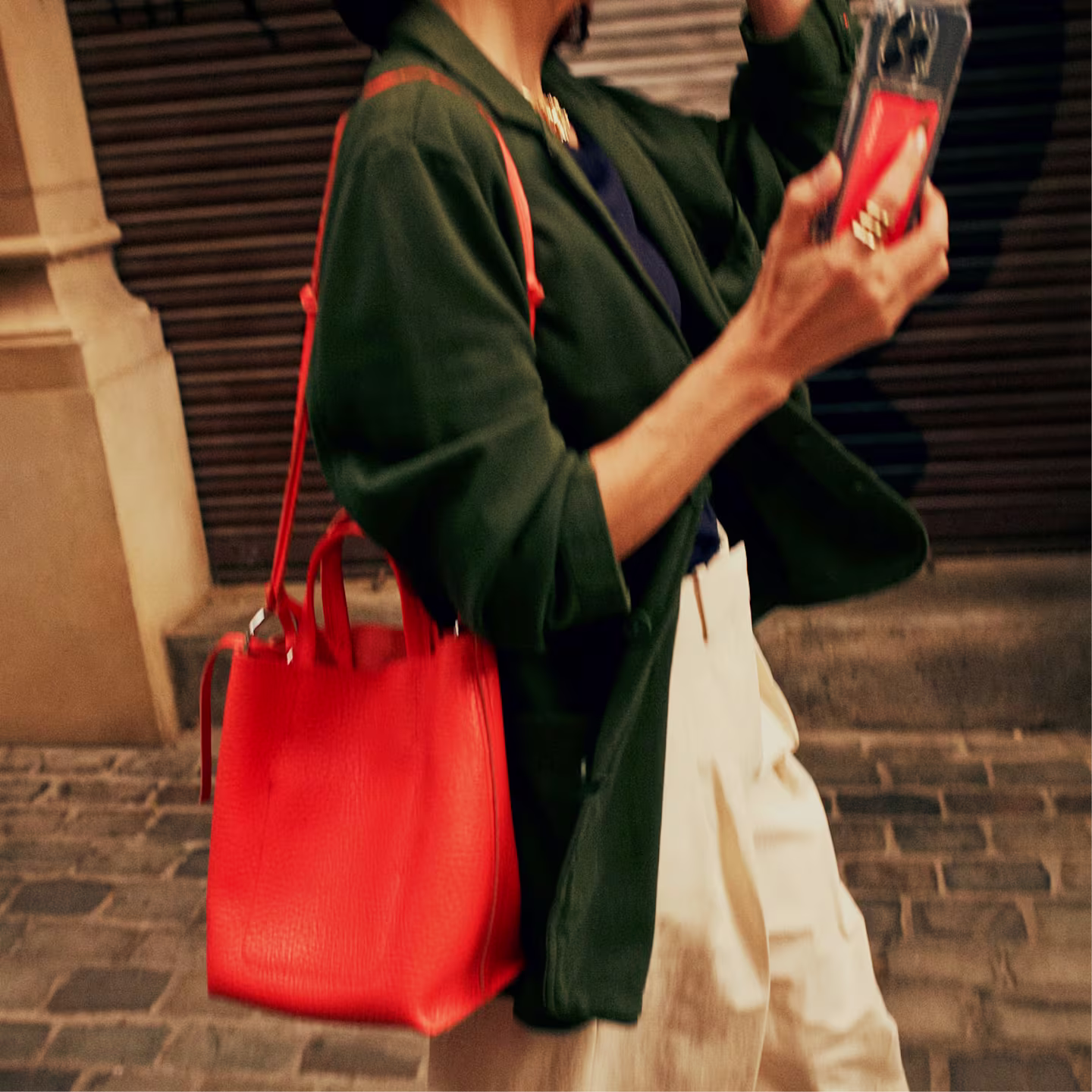 A person walks along a cobblestone street, wearing a dark green coat and cream trousers, carrying a hot coral coloured leather tote bag on their shoulder and holding a smartphone in their right hand. A Monzo debit card can be seen inside the phone case.