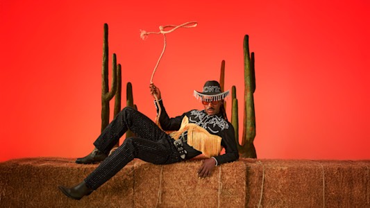 A person in a black cowboy hat, fringed shirt, and bedazzled pants reclines against a textured wall, holding a lasso. Two saguaro cacti stand behind them.