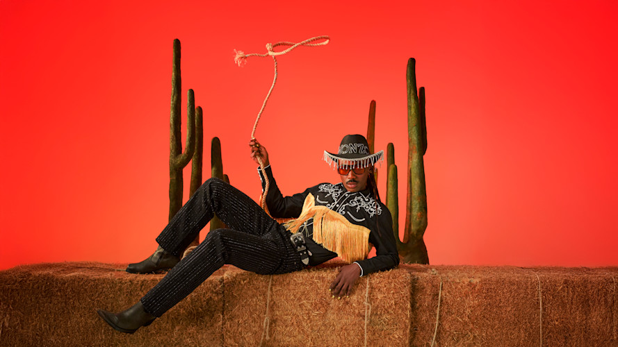 A person in a black cowboy hat, fringed shirt, and bedazzled pants reclines against a textured wall, holding a lasso. Two saguaro cacti stand behind them.