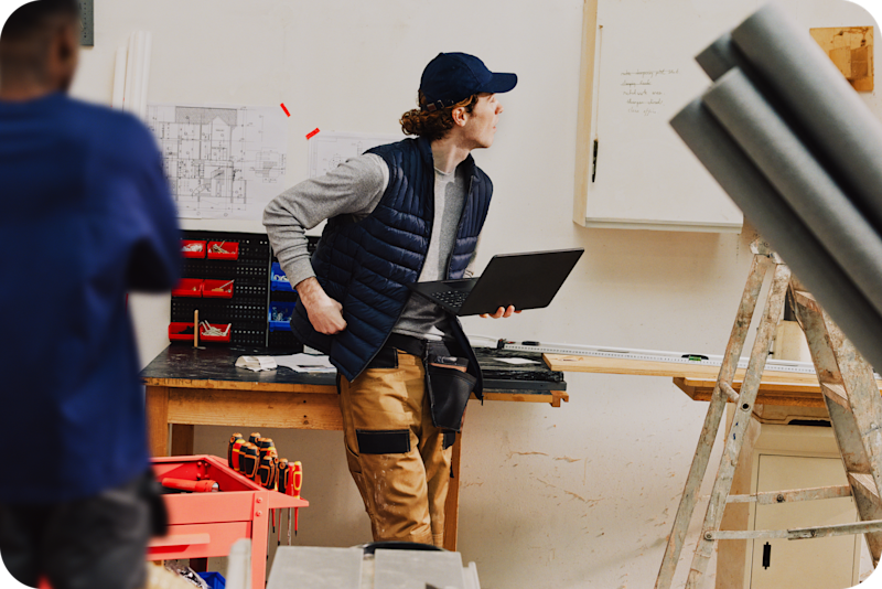 Construction workers collaborating on-site with a laptop and blueprints, one sitting on a workbench surrounded by tools ready to create quotes.