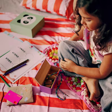 A young girl sitting on a striped bedspread with a pink Monzo debit card, craft supplies, and a jewelry box.