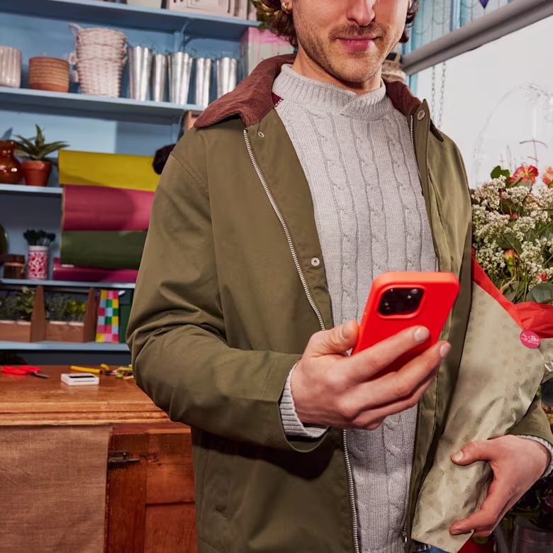 A man holds flowers in one hand and looks down at his hot coral phone in the other