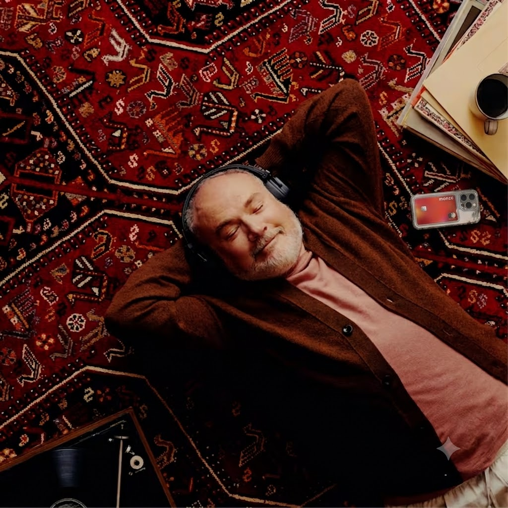 A man lays back on a deep red patterned rug. He rests his hands behind his head and closes his eyes. He has a relaxed smile and wears a pair of headphones.