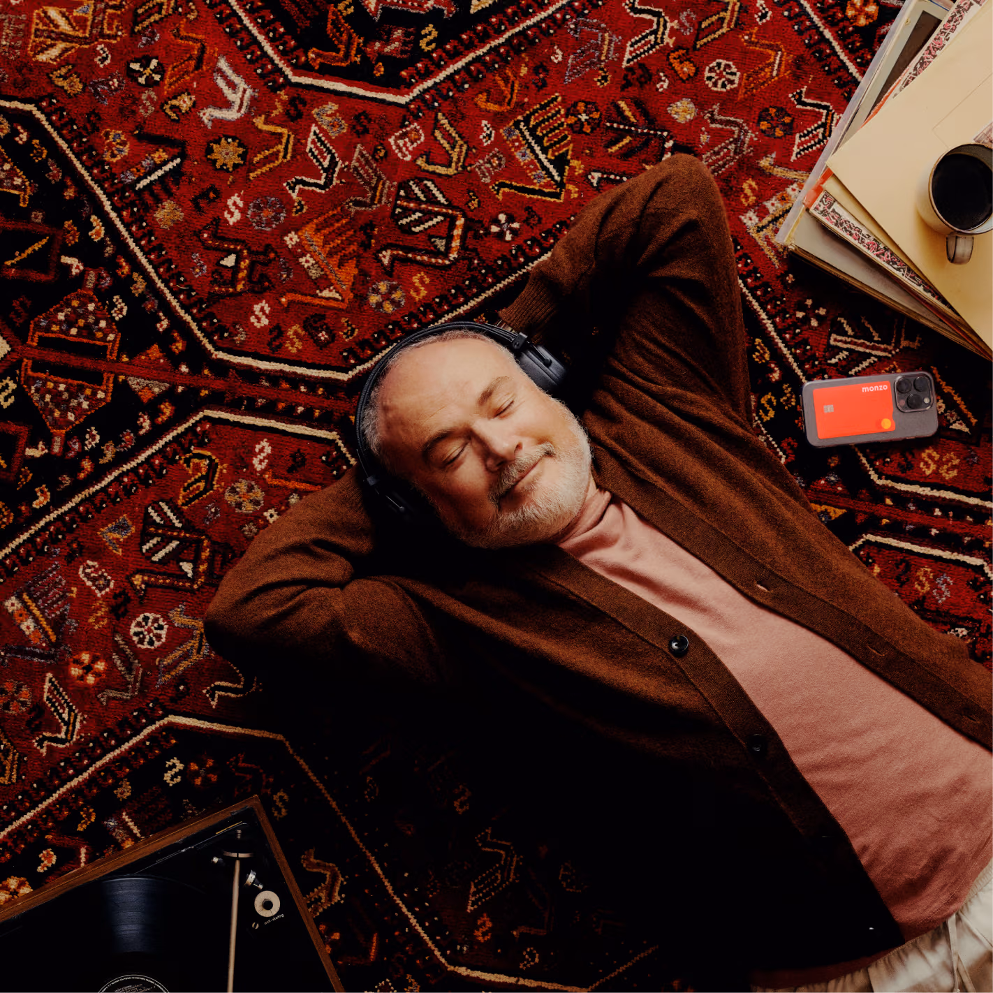 A man lays back on a deep red patterned rug. He rests his hands behind his head and closes his eyes. He has a relaxed smile and wears a pair of headphones.