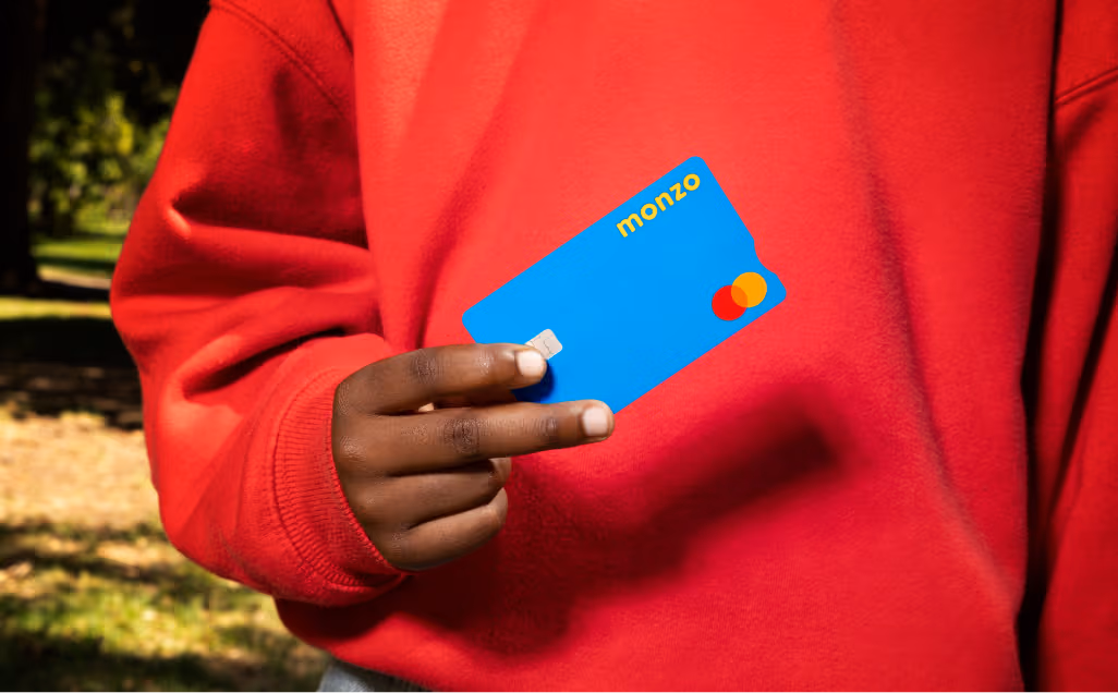 A close-up of a person wearing a bright red hoodie holding up a blue Monzo debit card.