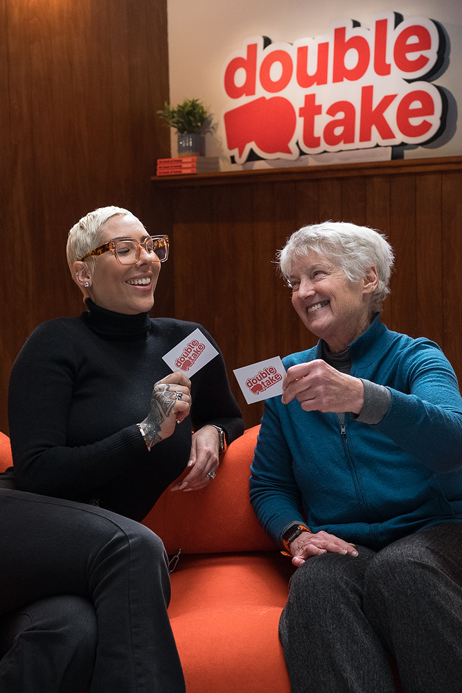 Two women sitting on a hot coral sofa chatting about investing, with a Double Take sign hanging in the background. 
