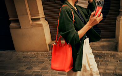 A person walking down the street with a large red handbag on their shoulder and their phone in their hand. You can see a Monzo card inside their phone case