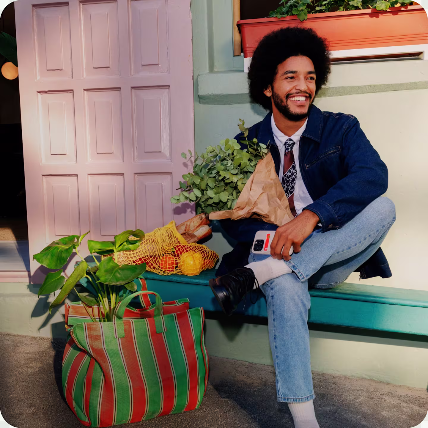 A smiling man with curly hair sits on a teal bench holding a bouquet of flowers next to a striped tote bag filled with fresh produce.