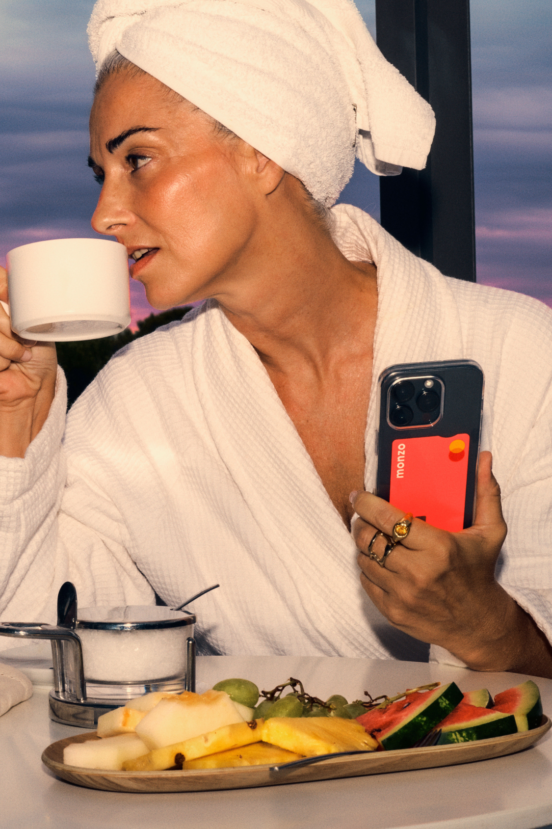 Photo of a woman at a hotel, sipping a cup of tea with a sunset in the background. 