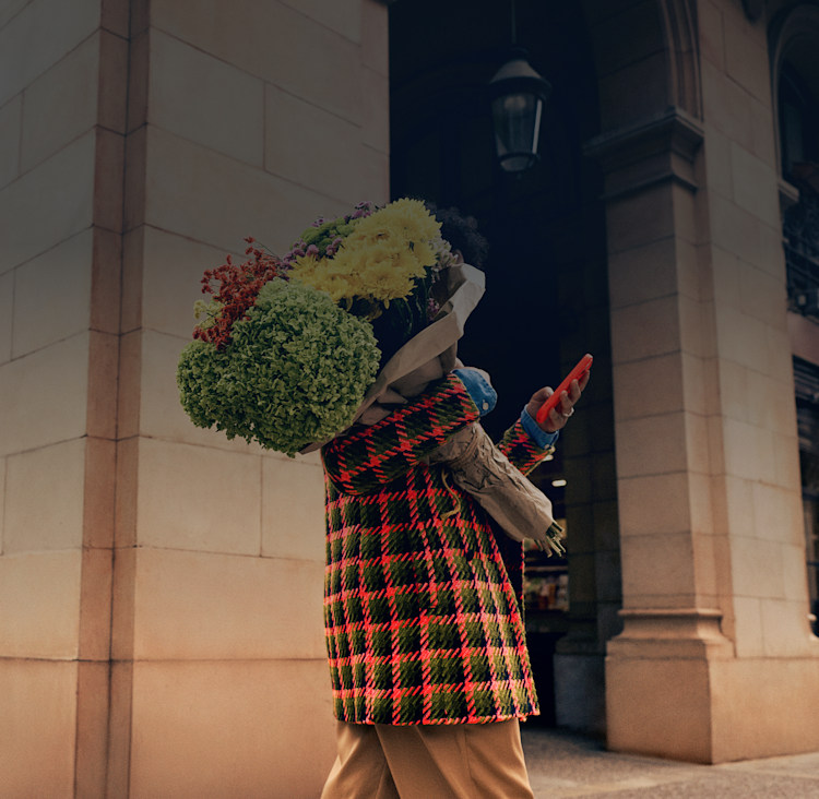 Person in plaid coat and tan pants stands near a building, holding a large bouquet of flowers and looking at the Monzo app on their phone.
