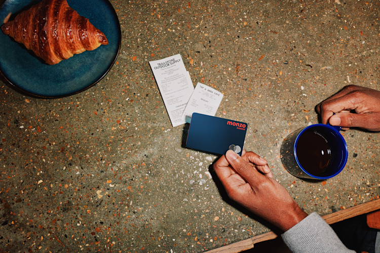 A business owner holds their Monzo Business card at a cafe counter with freshly baked croissant and coffee nearby.