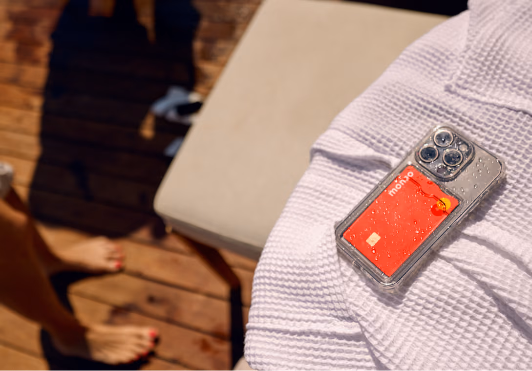 An aerial shot of mobile phone and Monzo debit card sitting on a white robe covering a sun lounger. A pair of sandals sit on a deck in the background.