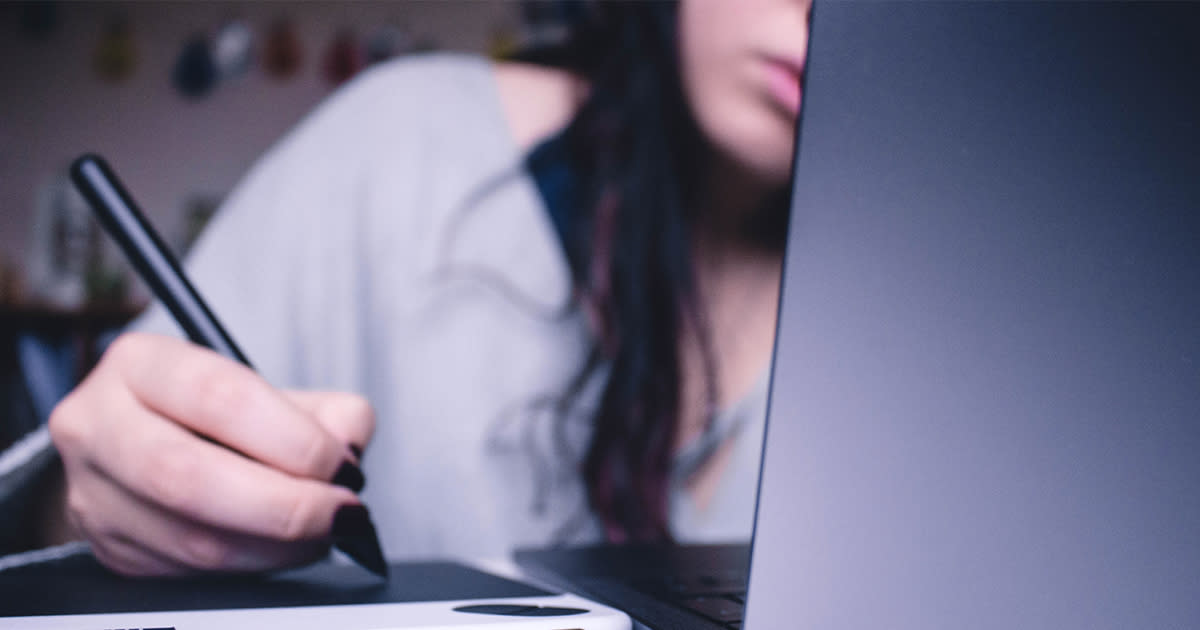 A person behind a laptop, appearing stressed, writing on a tablet