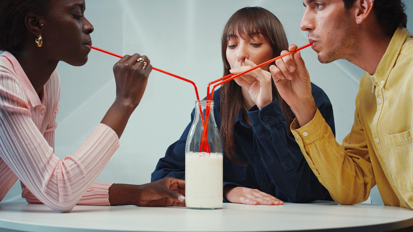 Three people share drinks from a single bottle using three red, multi-branched straws, sitting at a white table.