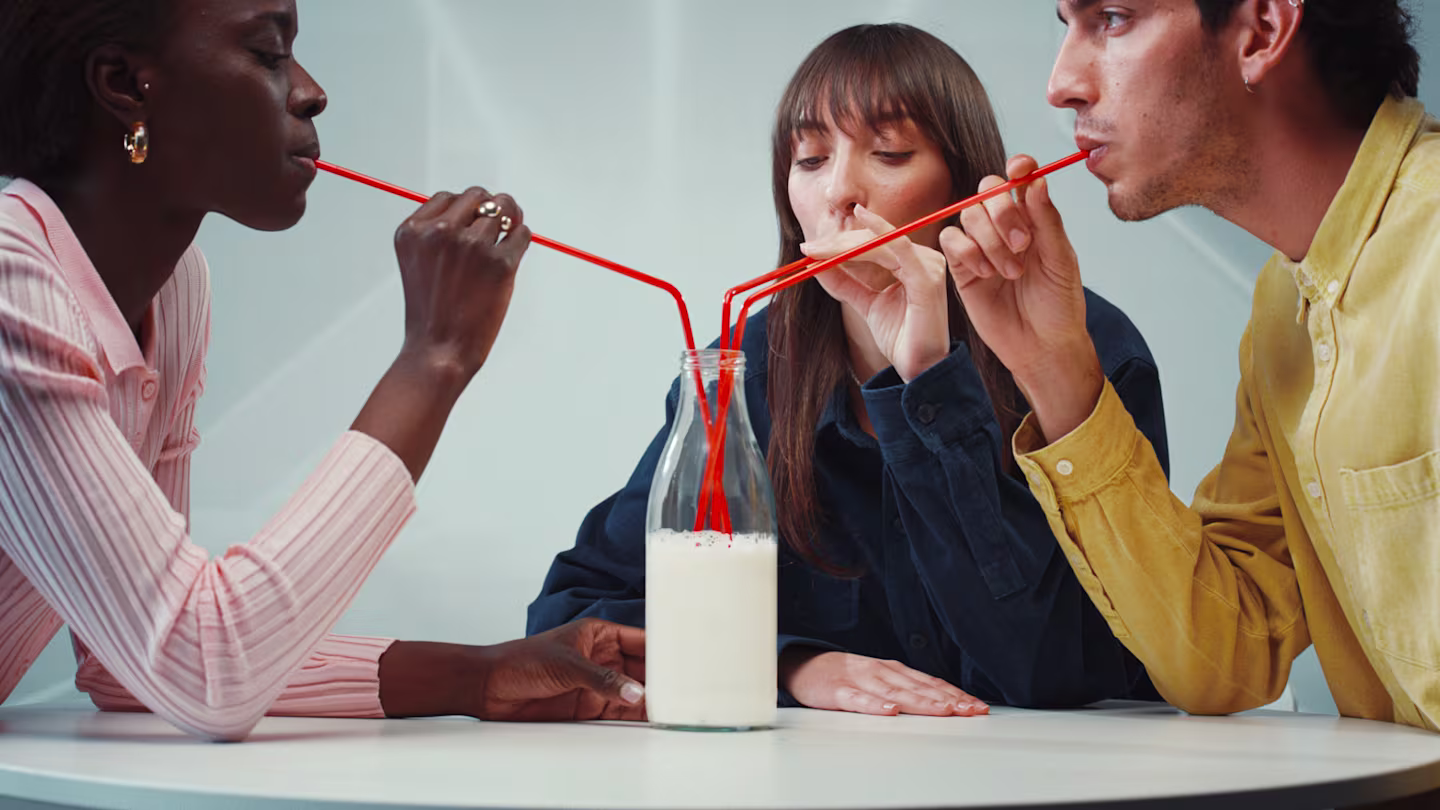 Three people share drinks from a single bottle using three red, multi-branched straws, sitting at a white table.