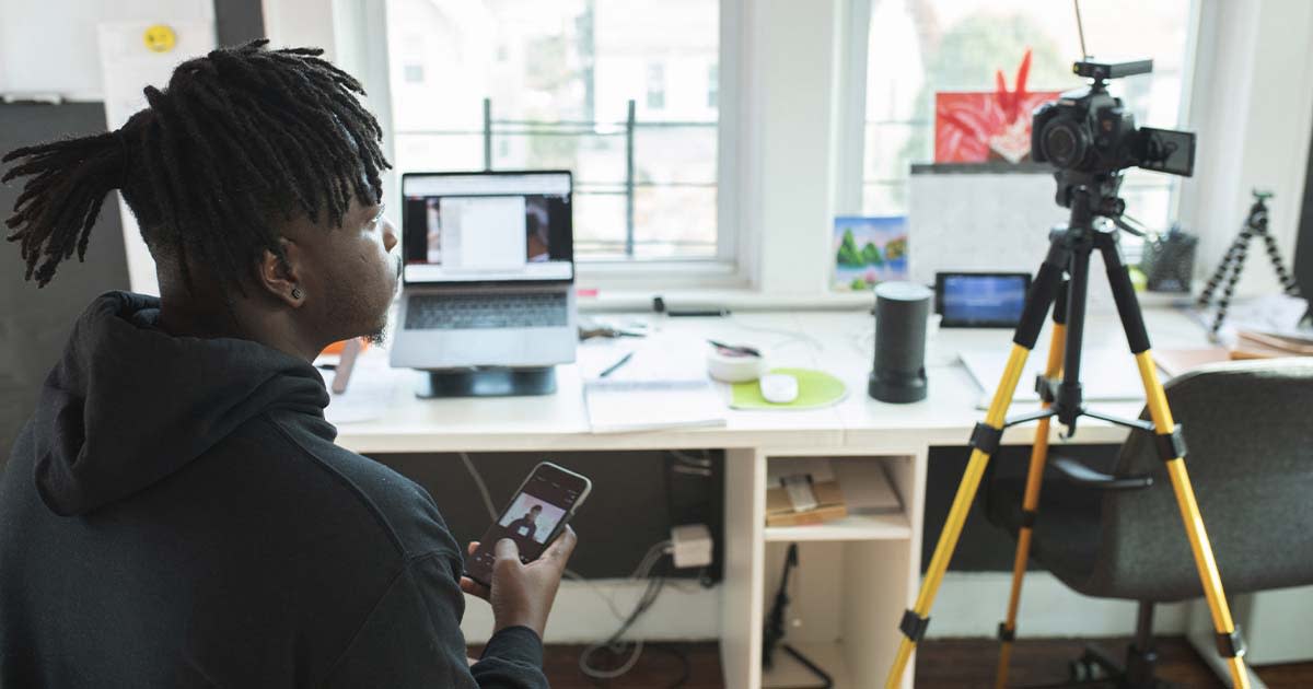 A man at home sitting by a desk with a laptop, holding a phone