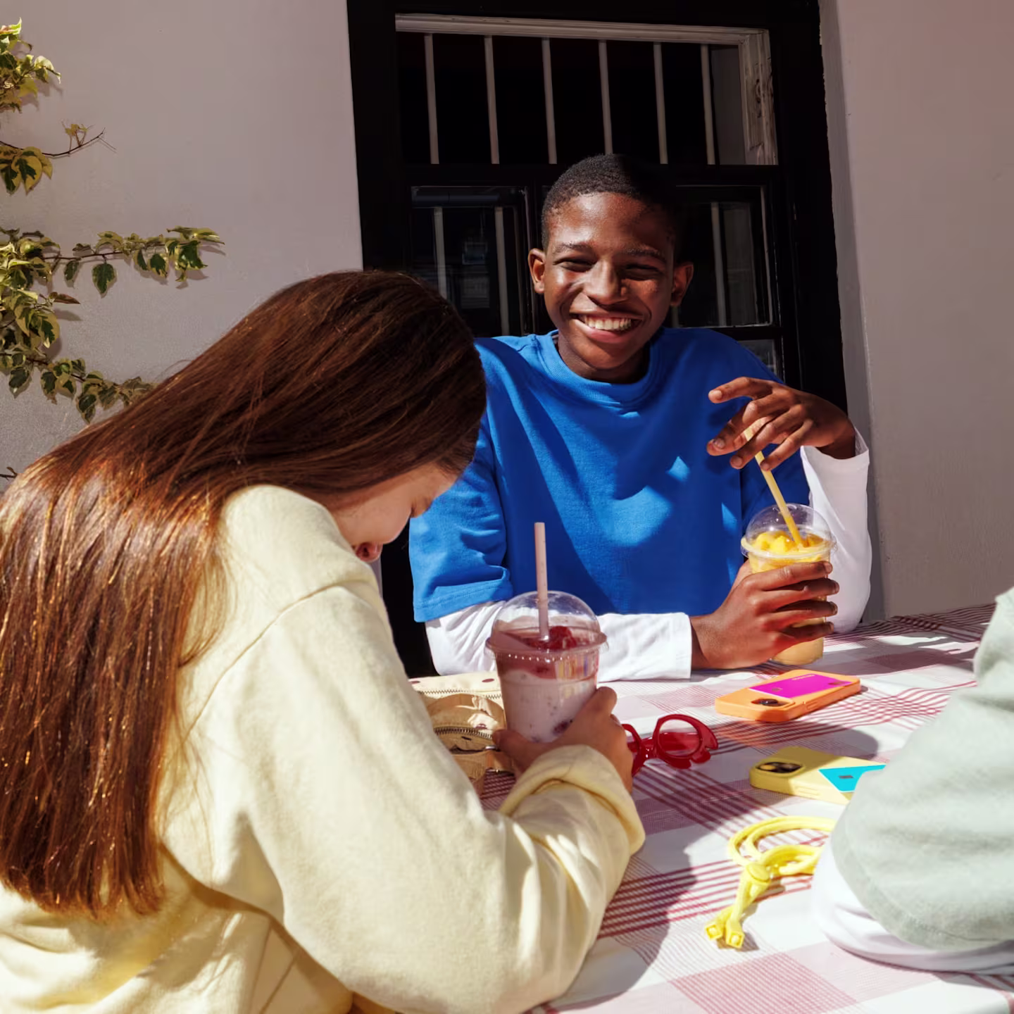 Three friends sitting at an outdoor table with drinks, chatting and smiling in the sunlight.