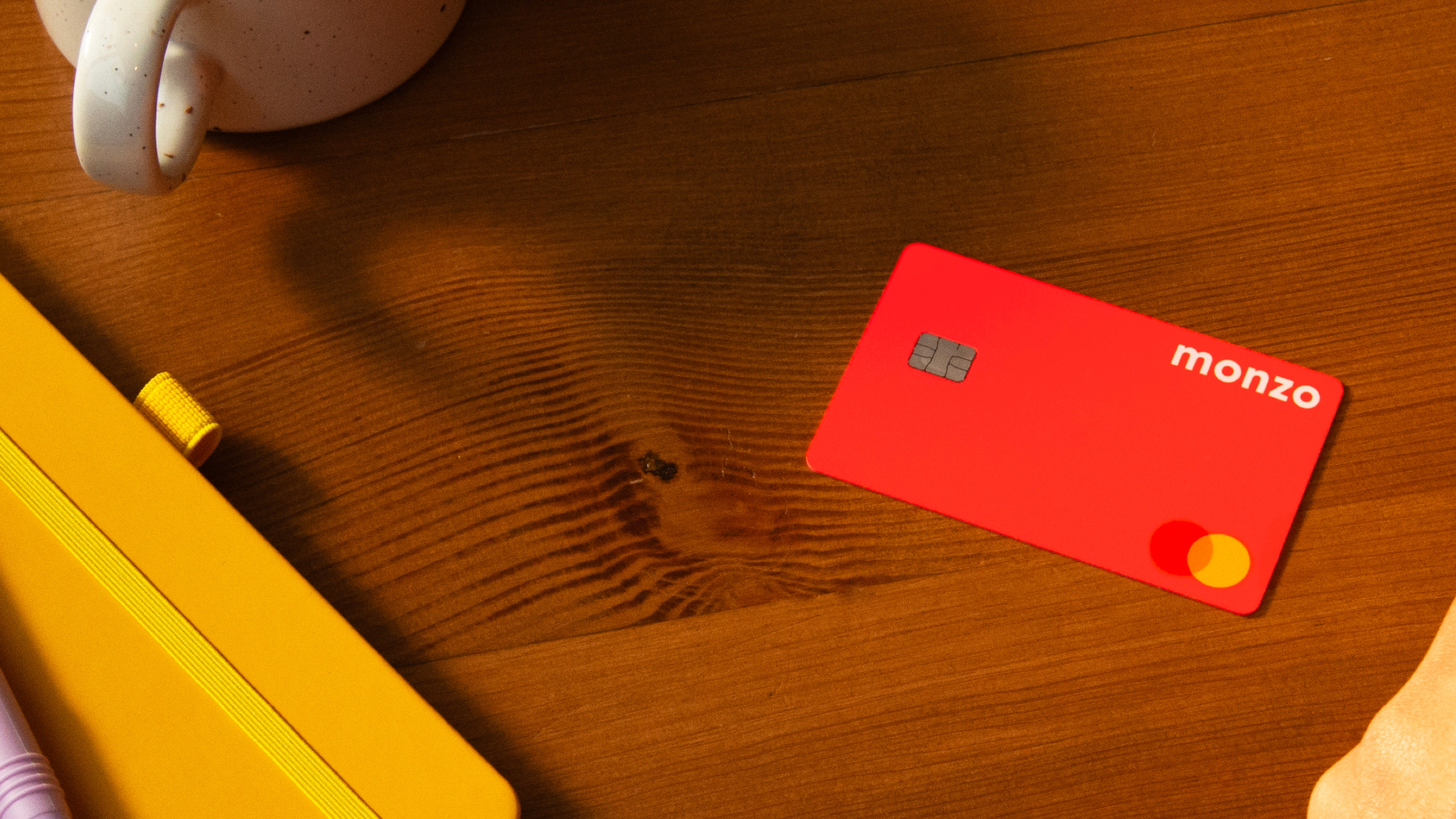 Monzo card sitting on brown wood table with a cup and notepad sitting off to the left side.