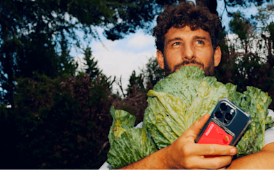 A close-up shot of a person with curly hair standing in a forest, holding a large head of leafy green lettuce up to their face. In their right hand, they hold a smartphone in a transparent case that reveals a bright red Monzo bank card tucked inside.