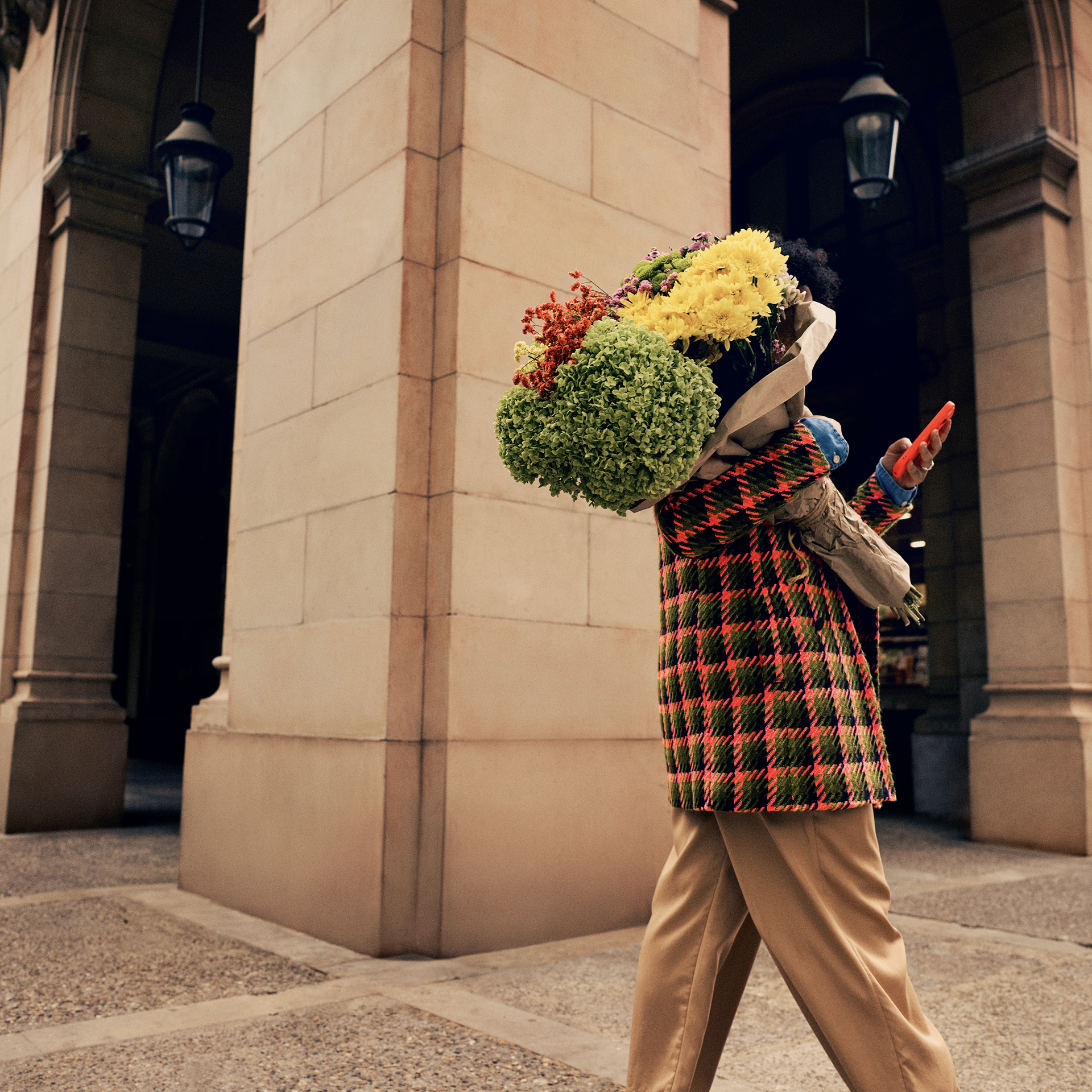 A lady in a Hot Coral and sage checkered coat is walking through a high street arcade. She has her back to us, but we can see the bright bouquet of flowers she carries over her shoulder as she checks her phone.