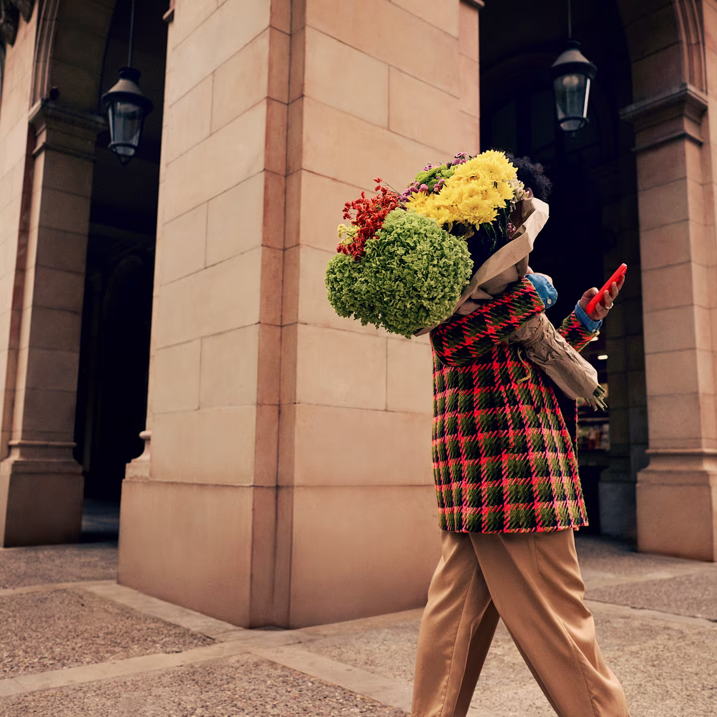 A lady in a Hot Coral and sage checkered coat is walking through a high street arcade. She has her back to us, but we can see the bright bouquet of flowers she carries over her shoulder as she checks her phone.