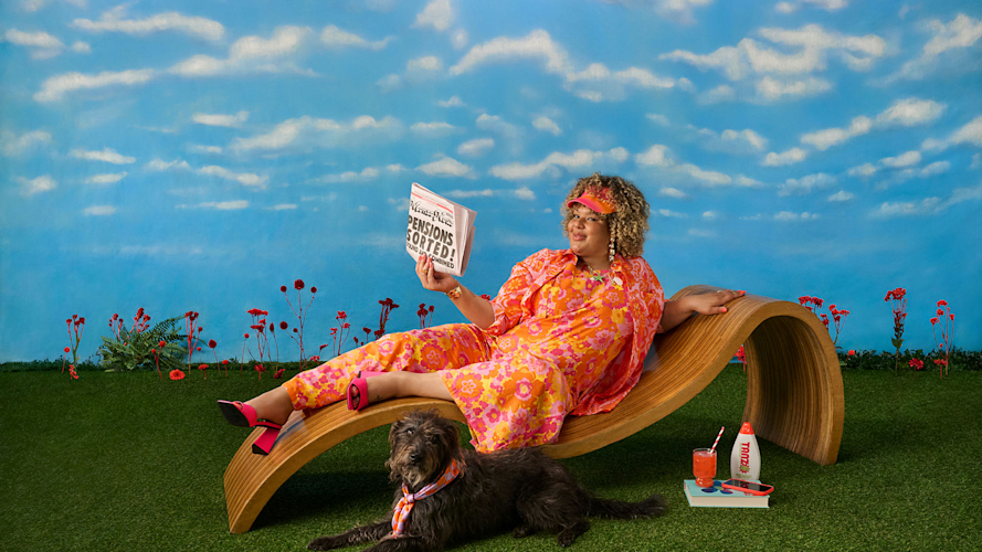 A person in an orange and pink floral outfit relaxes on a wooden lounge chair, reading a newspaper. A dog lies nearby.