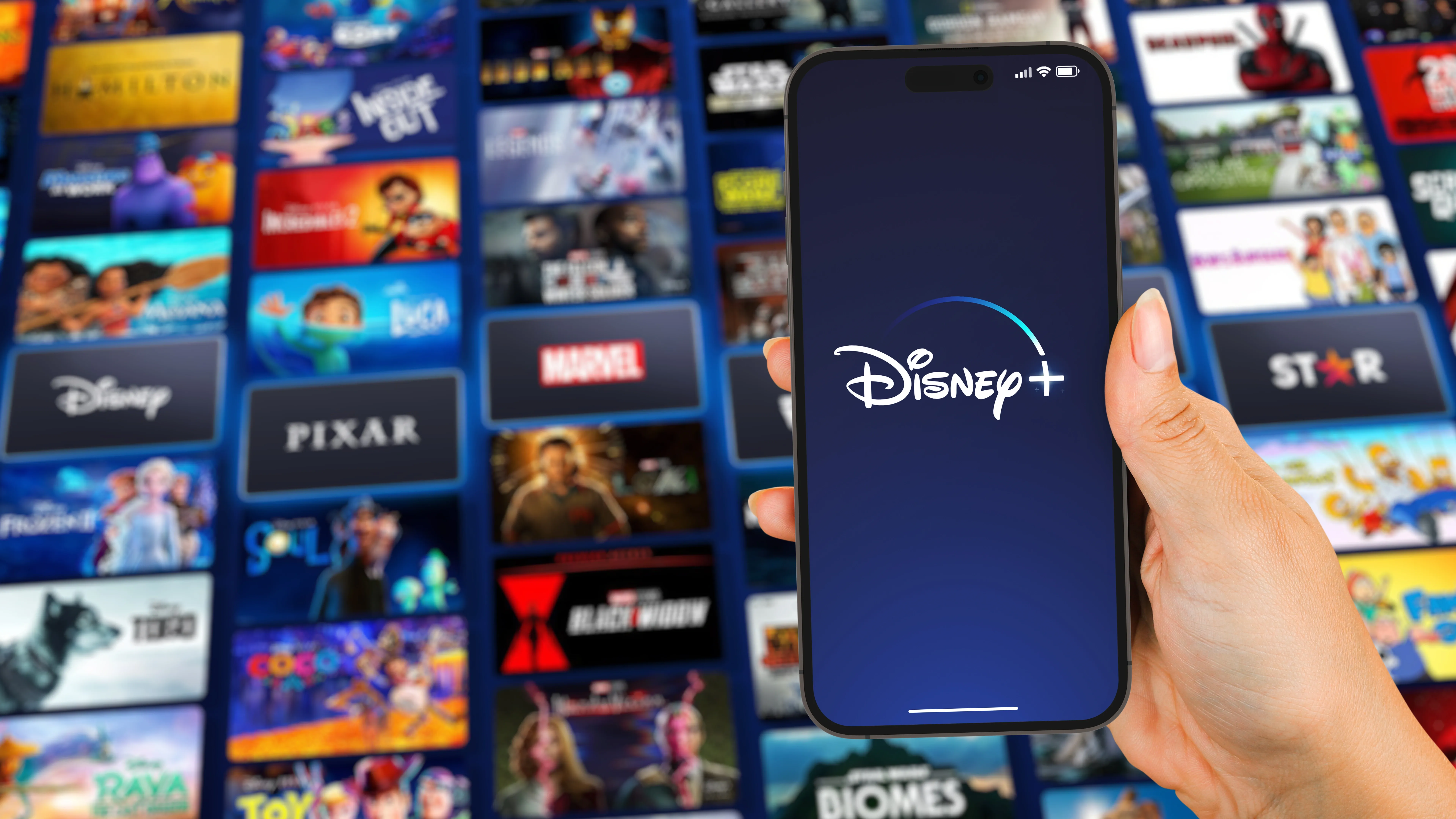 手に持ったスマホに映したDisney＋のロゴとその背後に並ぶ視聴できる作品群のサムネイル画像