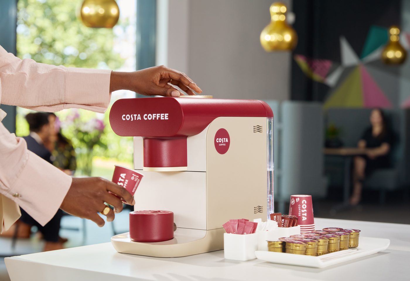 A person operates a Costa Coffee self-serve machine, placing a small Costa-branded cup under the dispenser. On the counter beside the machine are sugar packets, stirrers, and coffee pods, with people sitting in the background.