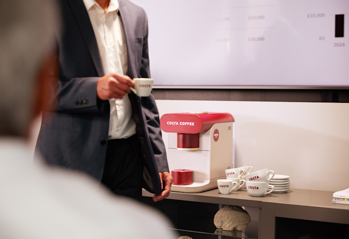 A person prepares a Costa Coffee using a coffee machine in a modern café setting, with coffee pods and condiments neatly arranged on the counter.