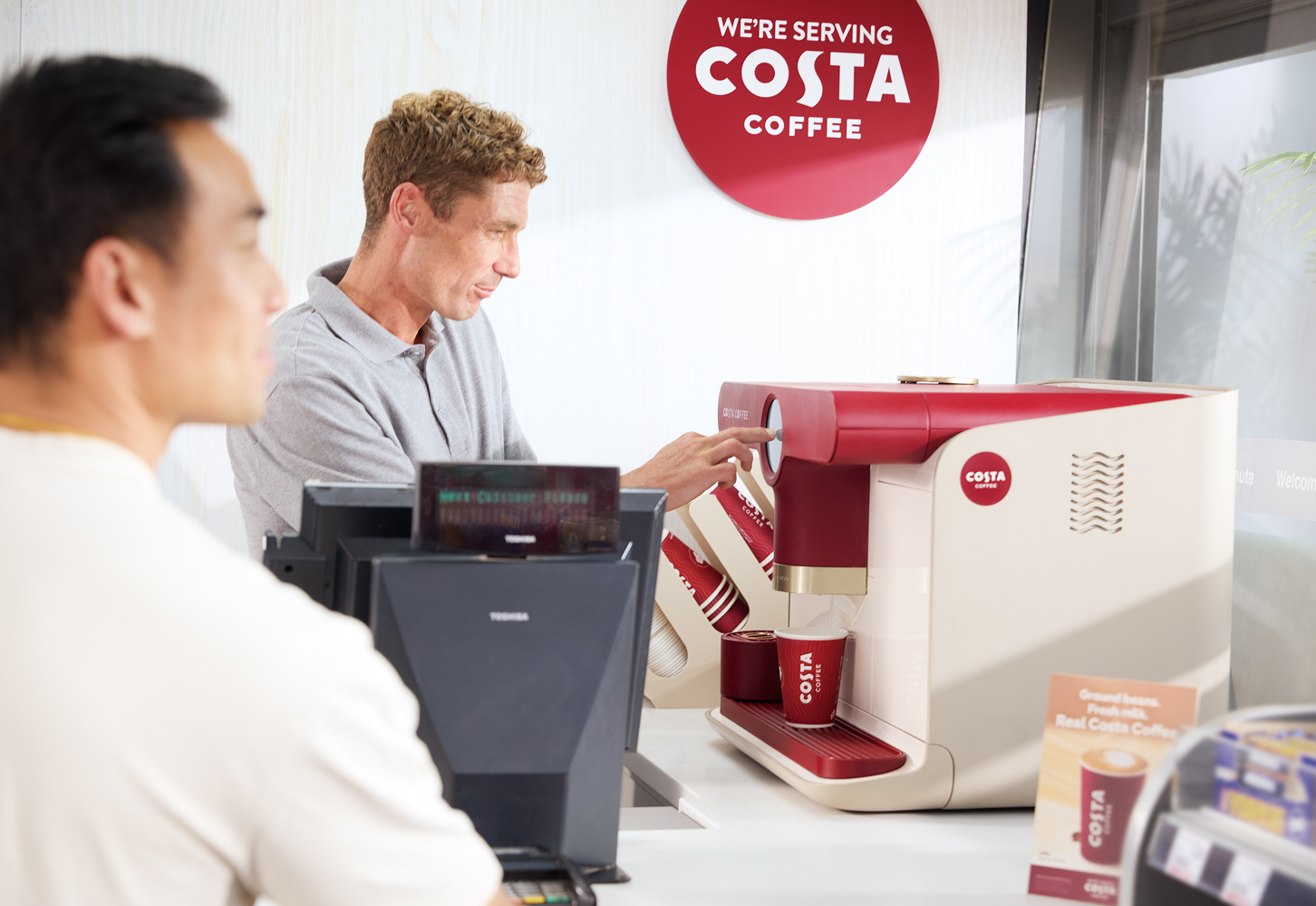 A man operates a red-and-white coffee machine at a café counter while another person waits nearby, with a “We’re serving Costa Coffee” sign visible on the wall.