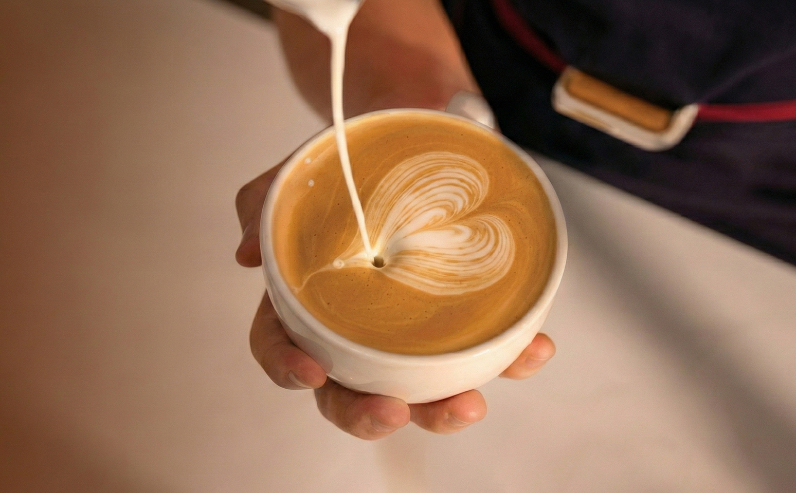 A barista pours steamed milk into a cup of coffee, creating a heart-shaped latte art design.