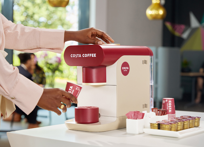 A person using a Costa Coffee self-serve coffee machine, placing a small Costa-branded paper cup under the dispenser, with sugar packets and coffee pods arranged on the counter nearby.