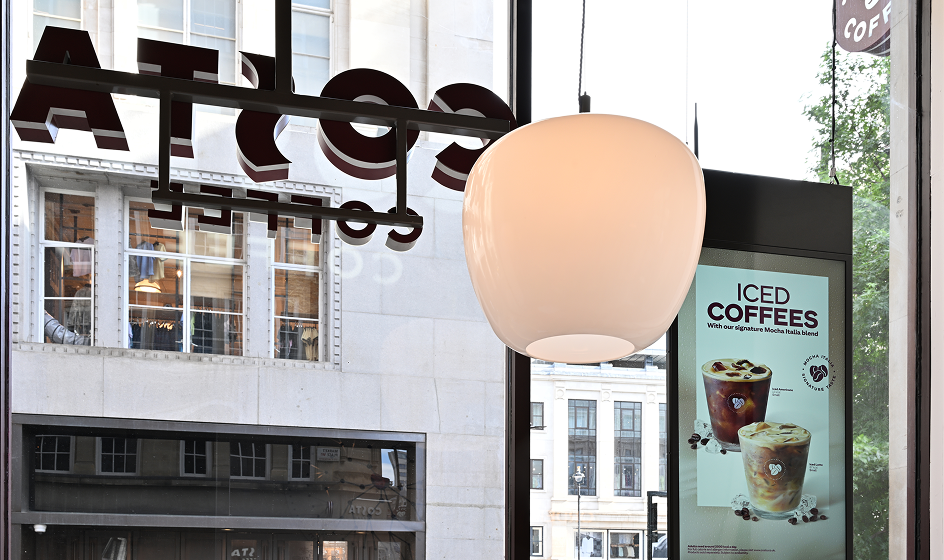 Interior view of a Costa Coffee café showing a hanging pendant light, a window with the Costa logo, and a digital sign advertising iced coffees, with city buildings outside.