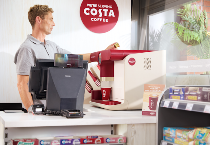 A man stands behind a counter operating a red-and-white Costa Coffee machine, with a cup placed under the dispenser and a “We’re serving Costa Coffee” sign on the wall.