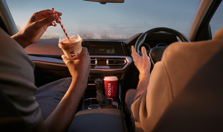 View from the back seat of a car showing two people in the front seats, one driving while the other holds an iced coffee with a striped straw; a takeaway coffee cup sits in the center console.