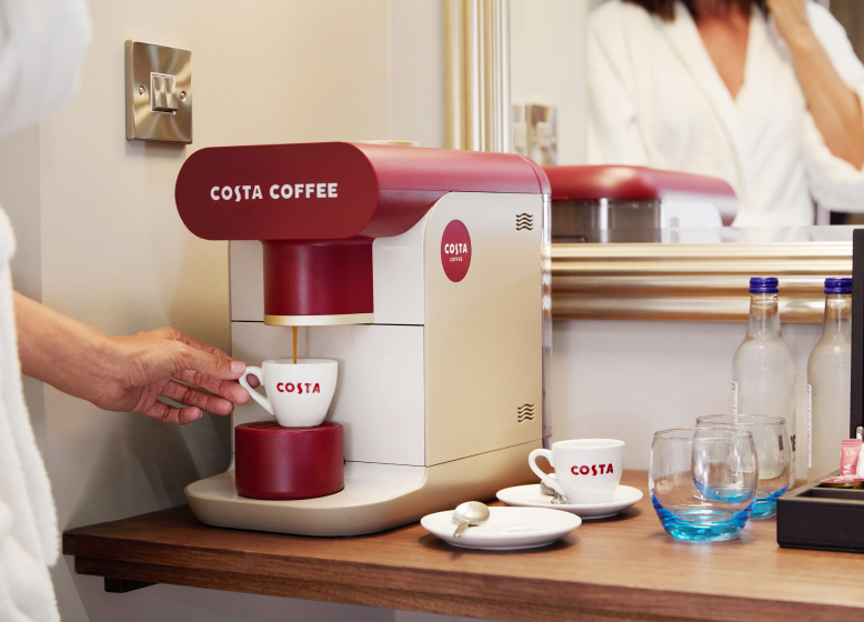 A person prepares a fresh cup of coffee using a Costa Coffee machine in a cozy setting, with water bottles, cups, and coffee essentials neatly arranged on the side