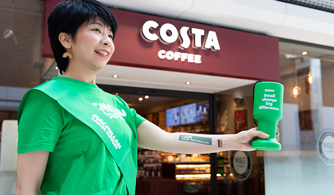 A woman in a green Macmillan shirt holding a green Macmillan donation cup in front of a Costa Coffee store