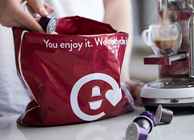 Hand drops a used coffee capsule into a maroon recycling bag labeled “You enjoy it. We recycle,” beside an espresso machine.