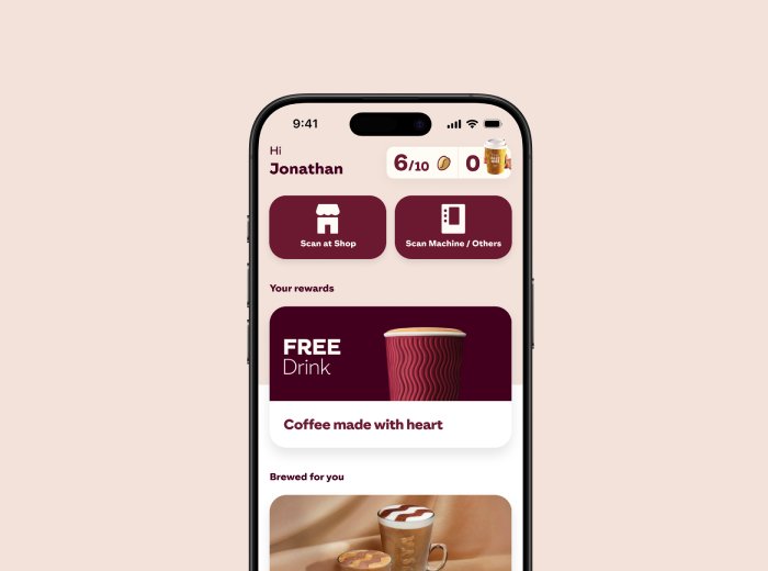 A smartphone screen showing a coffee shop rewards app. The top of the screen displays a greeting "Hi Jonathan" with a progress indicator showing "6/10" stamps, "0" coins, and a drink icon. Below are two buttons: "Scan at Shop" and "Scan Machine/Others." The "Your rewards" section shows a reward for a "FREE Drink" with an image of a takeaway coffee cup. Underneath, text reads "Coffee made with heart." The lower part of the screen shows a "Brewed for you" section with an image of a coffee cup.