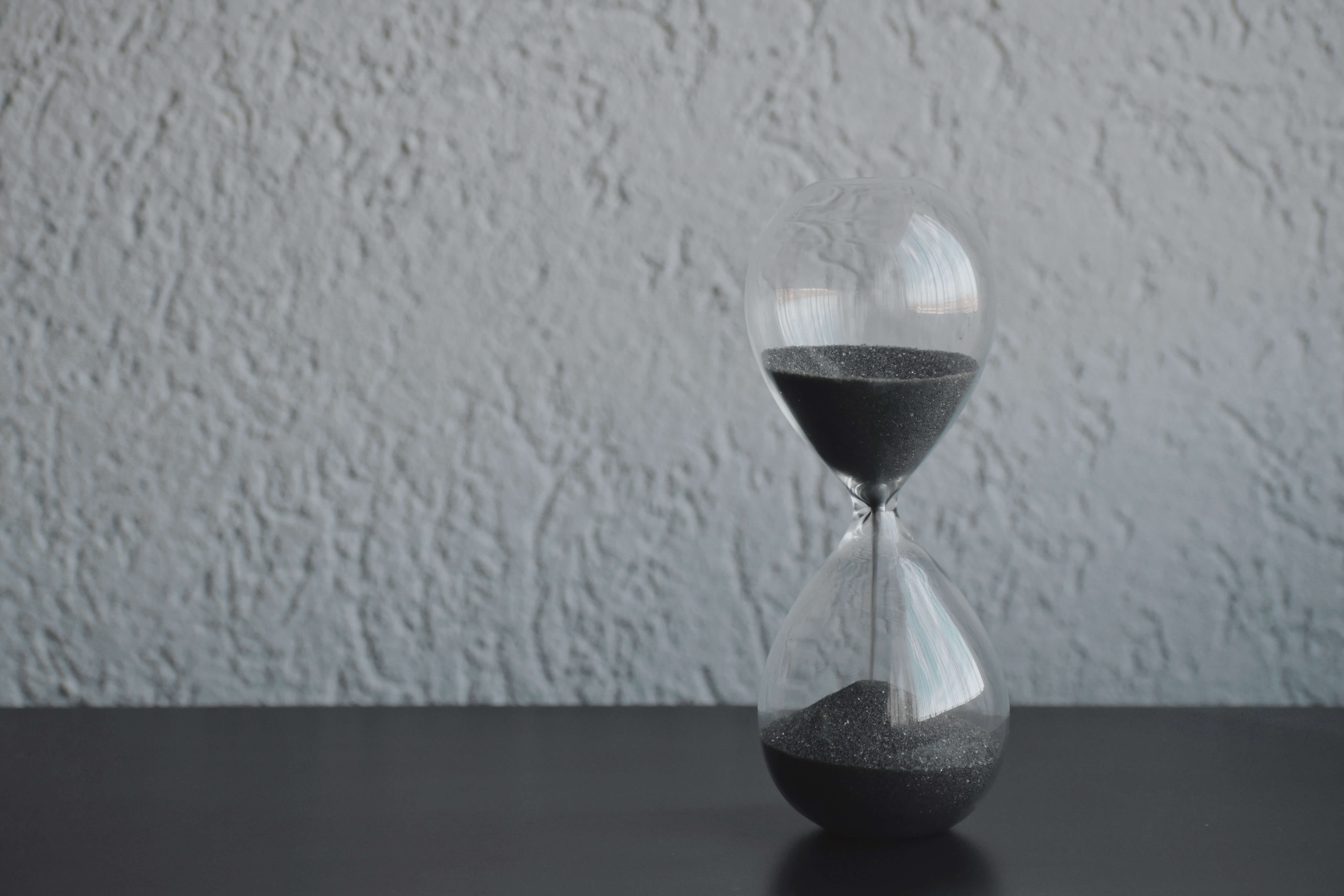 An image of an hour glass with black sand, on a black surface, against a white wall