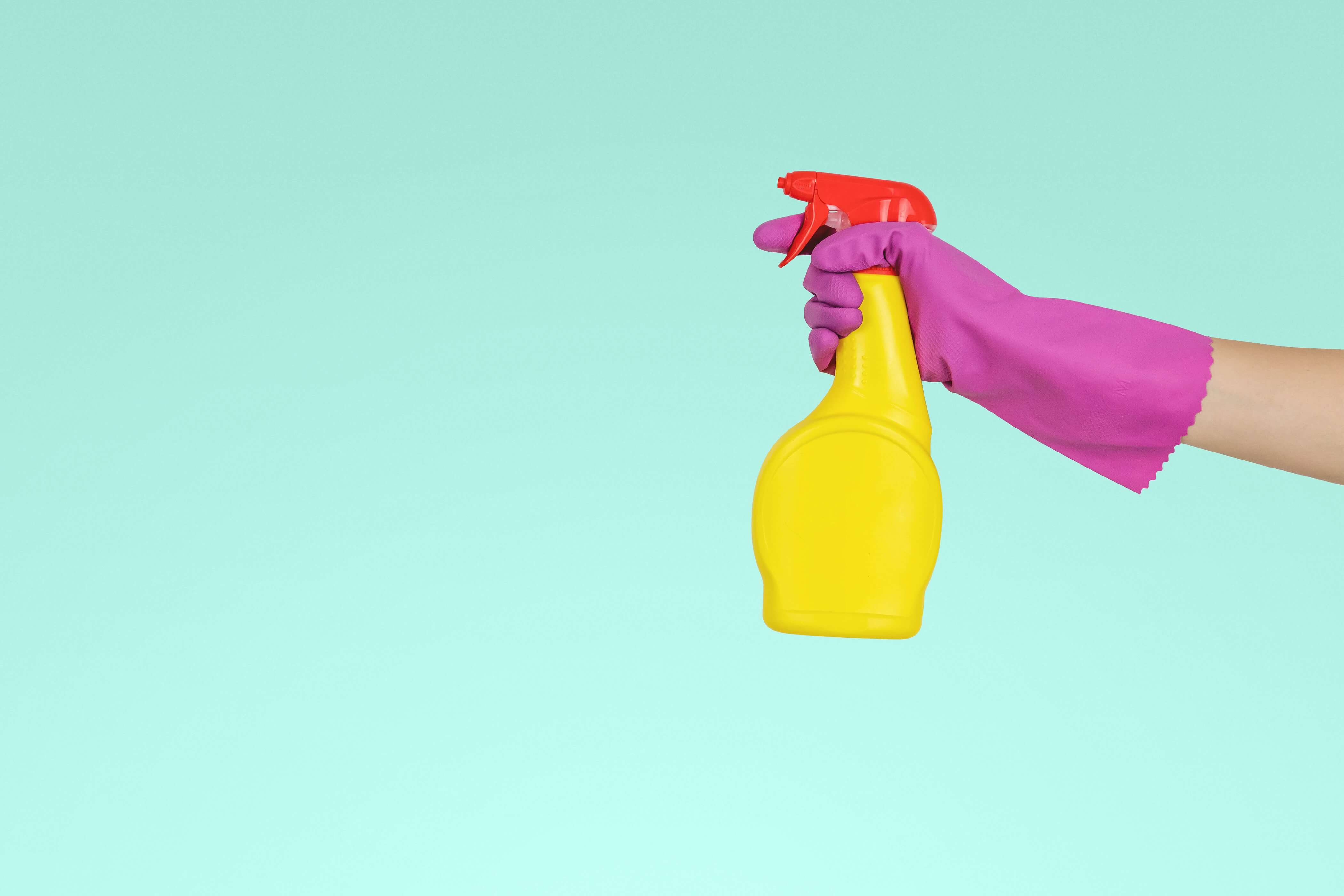 Someones hand in a pink rubber glove, holding a yellow spray bottle with cleaning products 