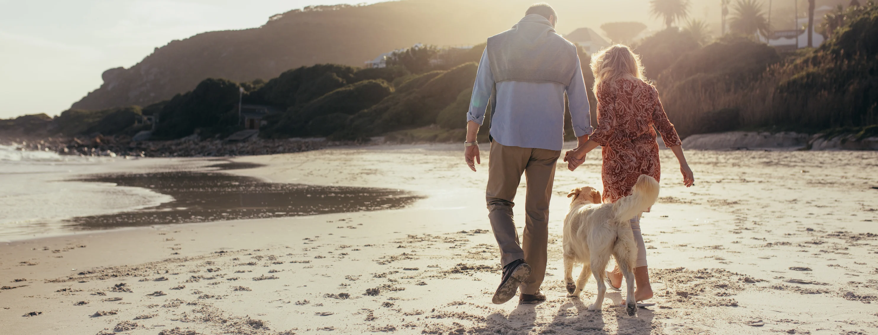 A man and woman hold hands on the beach with their golden retriever following them