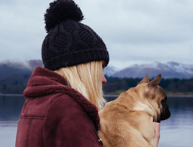 Blonde woman with a black beanie wearing a red jacket is holding her pug dog on a boat dock in winter.