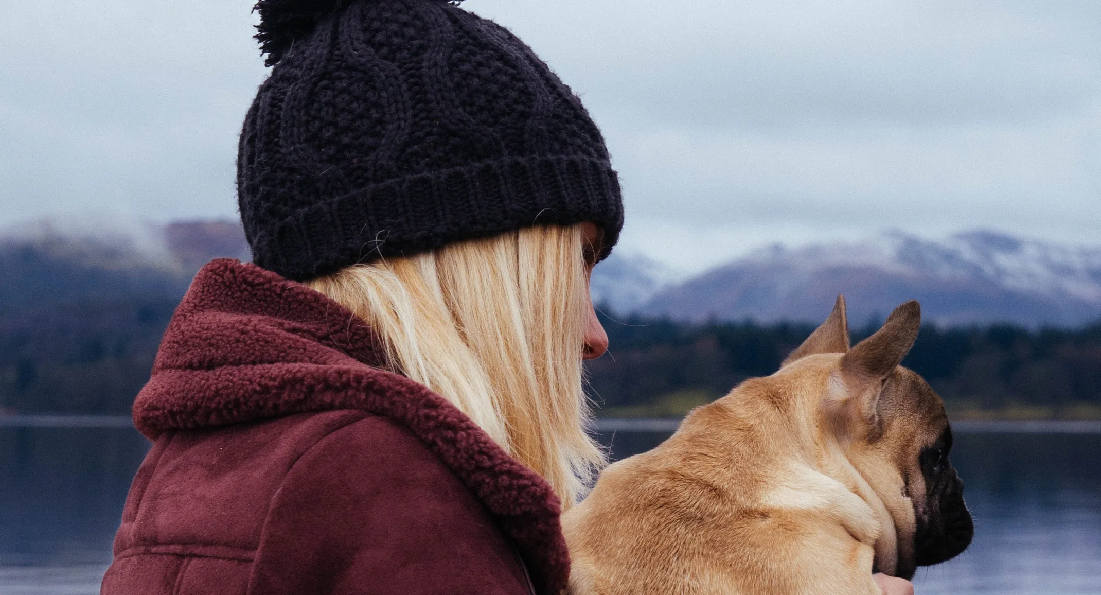 Blonde woman with a black beanie wearing a red jacket is holding her pug dog on a boat dock in winter.