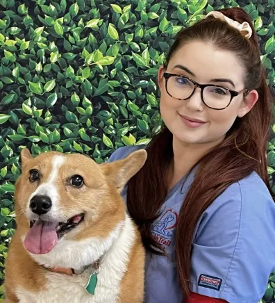 Marina's staff photo from Spanish Trail Animal Hospital where she is outside sitting on a lawn posing for a picture next to tan and white corgi dog.