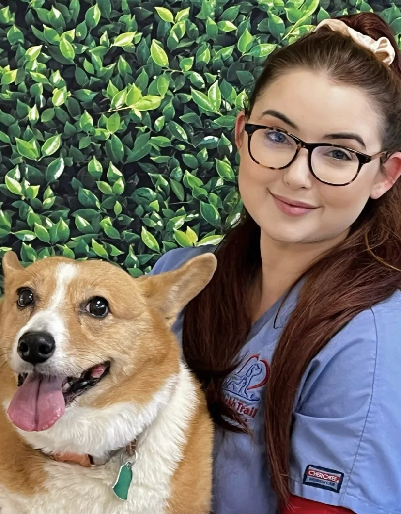 Marina's staff photo from Spanish Trail Animal Hospital where she is outside sitting on a lawn posing for a picture next to tan and white corgi dog.