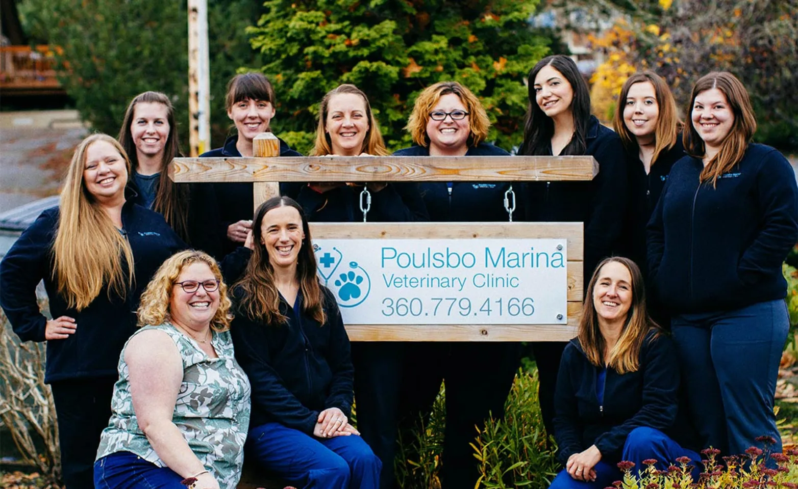 Poulsbo Marina Veterinary Clinic is posing for a group photo outside and next to their clinic's sign.  There's eleven women in the picture. 